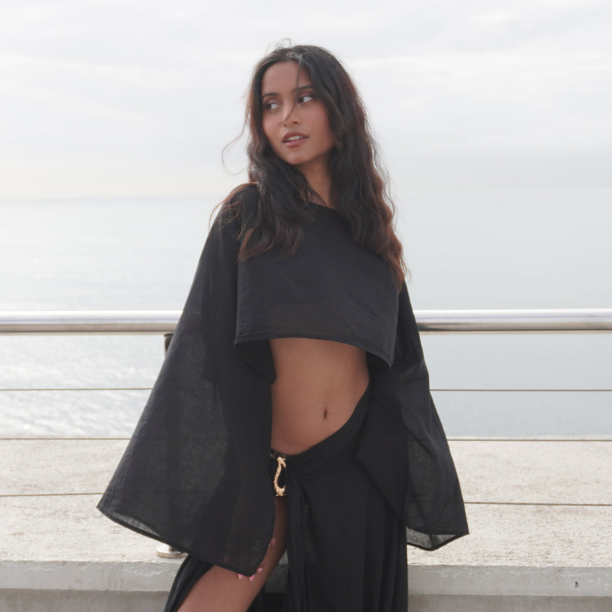 A woman wearing a black two-piece outfit with a cropped long-sleeve top and matching draped skirt stands outdoors near a railing with water in the background. The Phoenix set, sheer black cotton beachwear by Nonza, made in France.