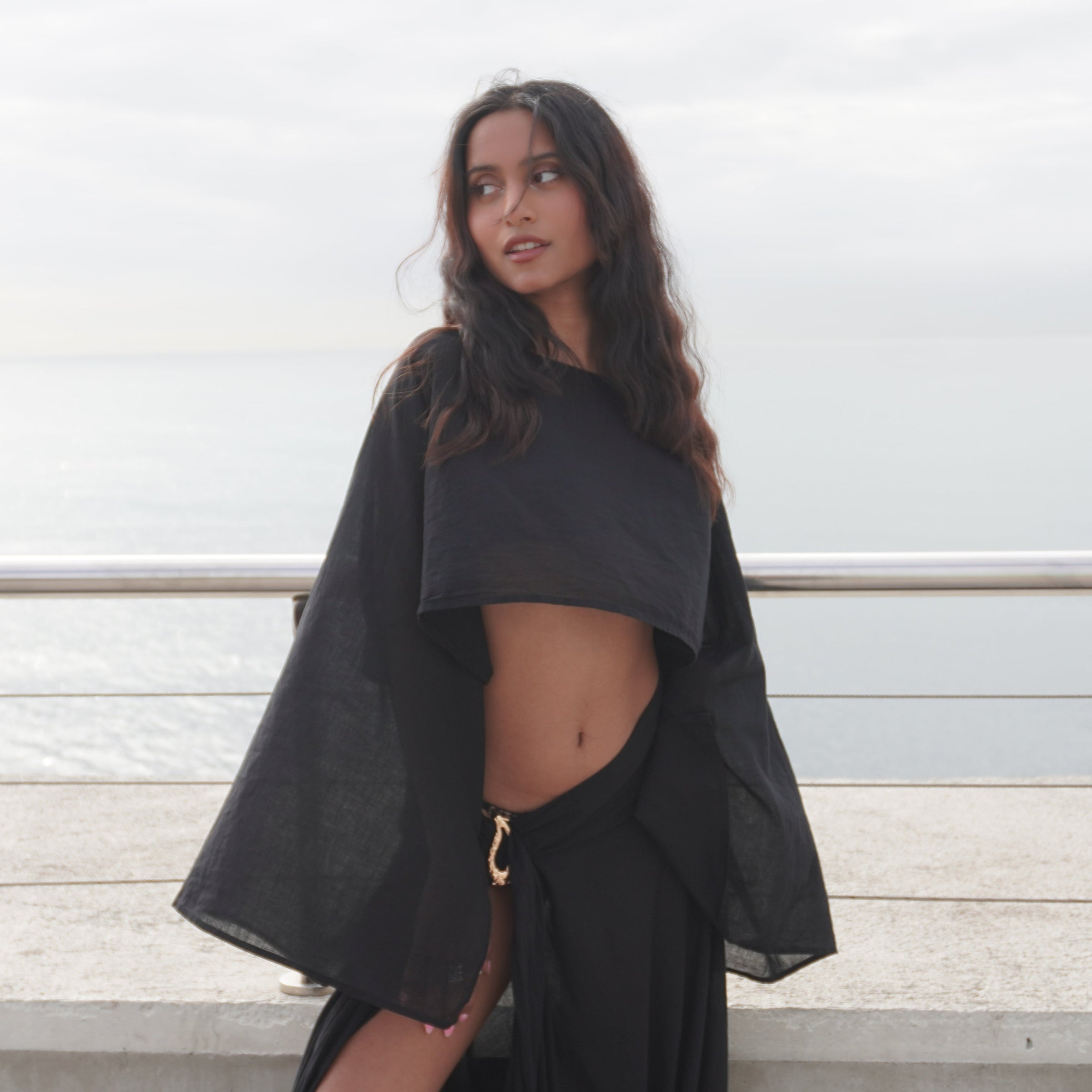 A woman wearing a black two-piece outfit with a cropped long-sleeve top and matching draped skirt stands outdoors near a railing with water in the background. The Phoenix set, sheer black cotton beachwear by Nonza, made in France.