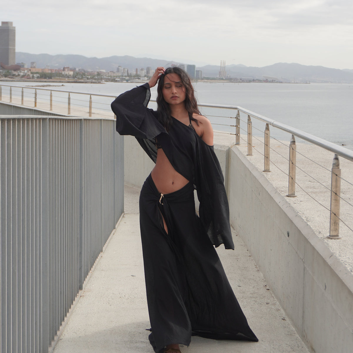 A woman wearing a black long-sleeve crop top and matching wide-leg pants poses on an outdoor concrete walkway overlooking water. The Phoenix set, sheer black cotton beachwear by Nonza, made in France.